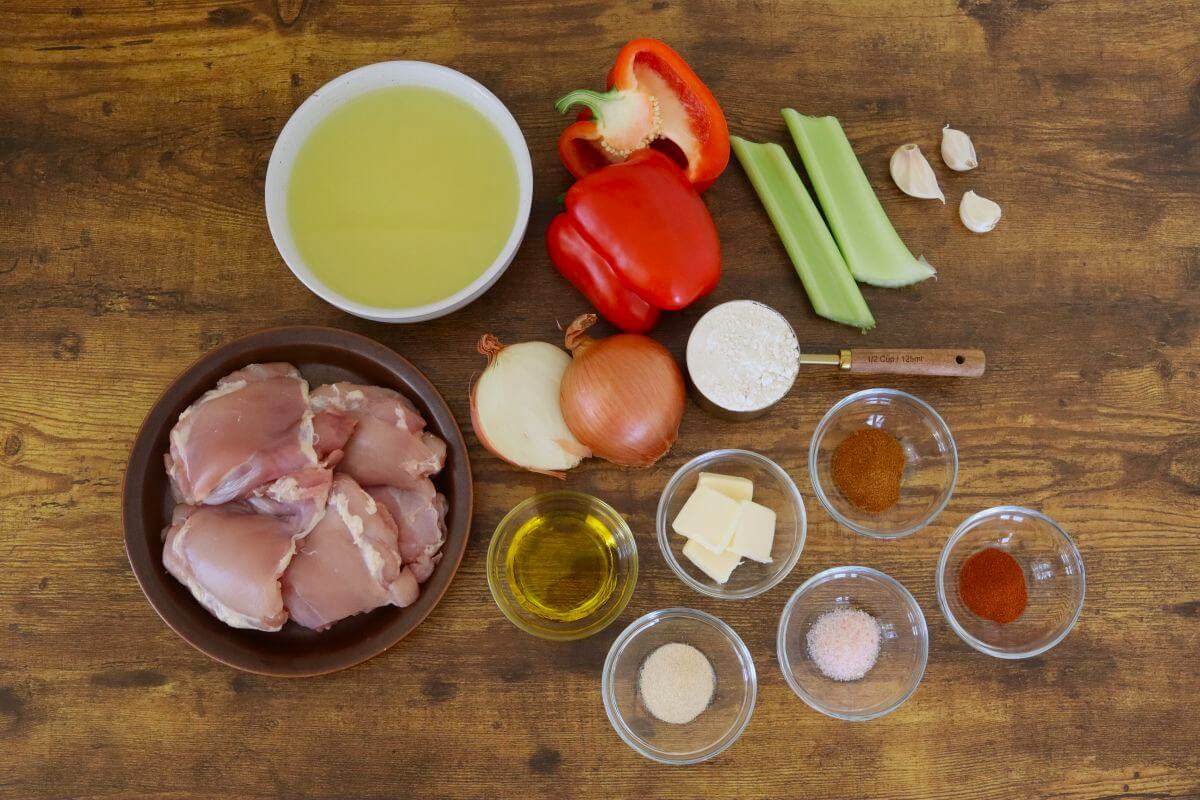 Raw chicken thighs, chicken broth, bell pepper, onion, celery, garlic, flour, butter, oil, and spices arranged on a wooden surface.