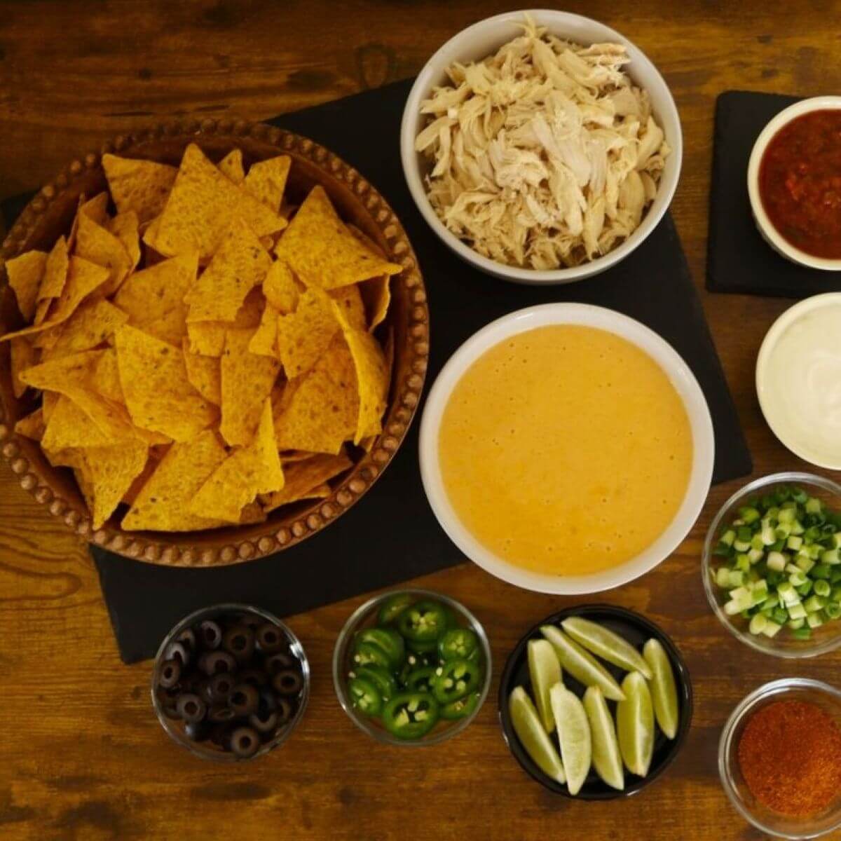 Tortilla chips, shredded chicken, cheese sauce, olives, jalapeños, lime wedges, and scallions in bowls on a table.