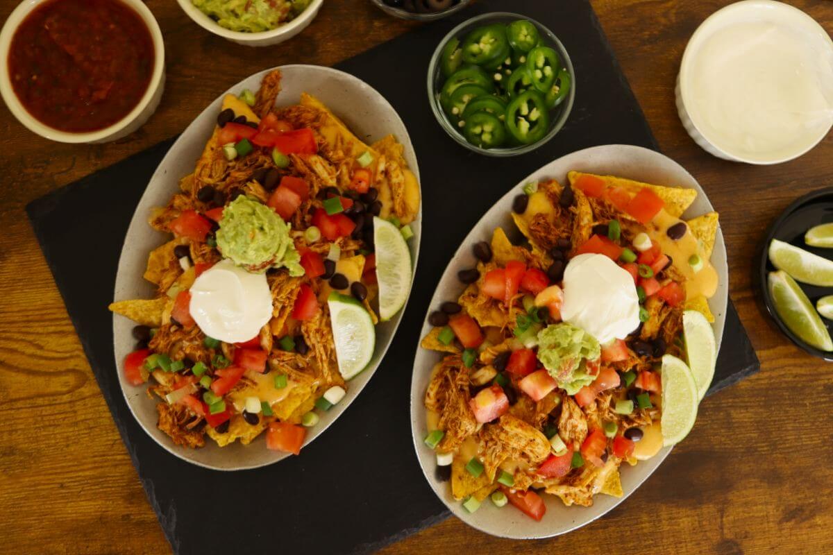 Two plates of chicken nachos with toppings, surrounded by bowls of salsa, jalapeños, and lime wedges.