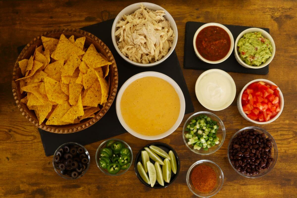 Tortilla chips, shredded chicken, cheese sauce, and assorted nacho toppings arranged in bowls on a wooden table.