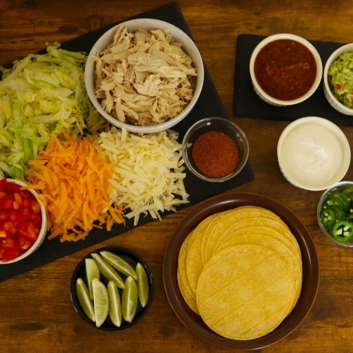 Shredded chicken, shredded lettuce, cheddar, Monterey jack, diced tomatoes, tortillas, lime wedges, salsa, guacamole, sour cream, and jalapeños on wooden table.