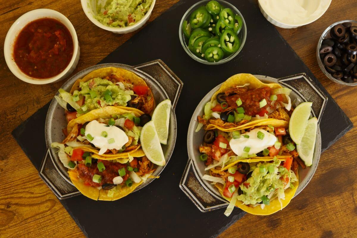 Two plates of shredded chicken tacos with lettuce, tomatoes, cheese, salsa, sour cream, and lime wedges beside small bowls of guacamole, jalapeños, olives, and salsa.