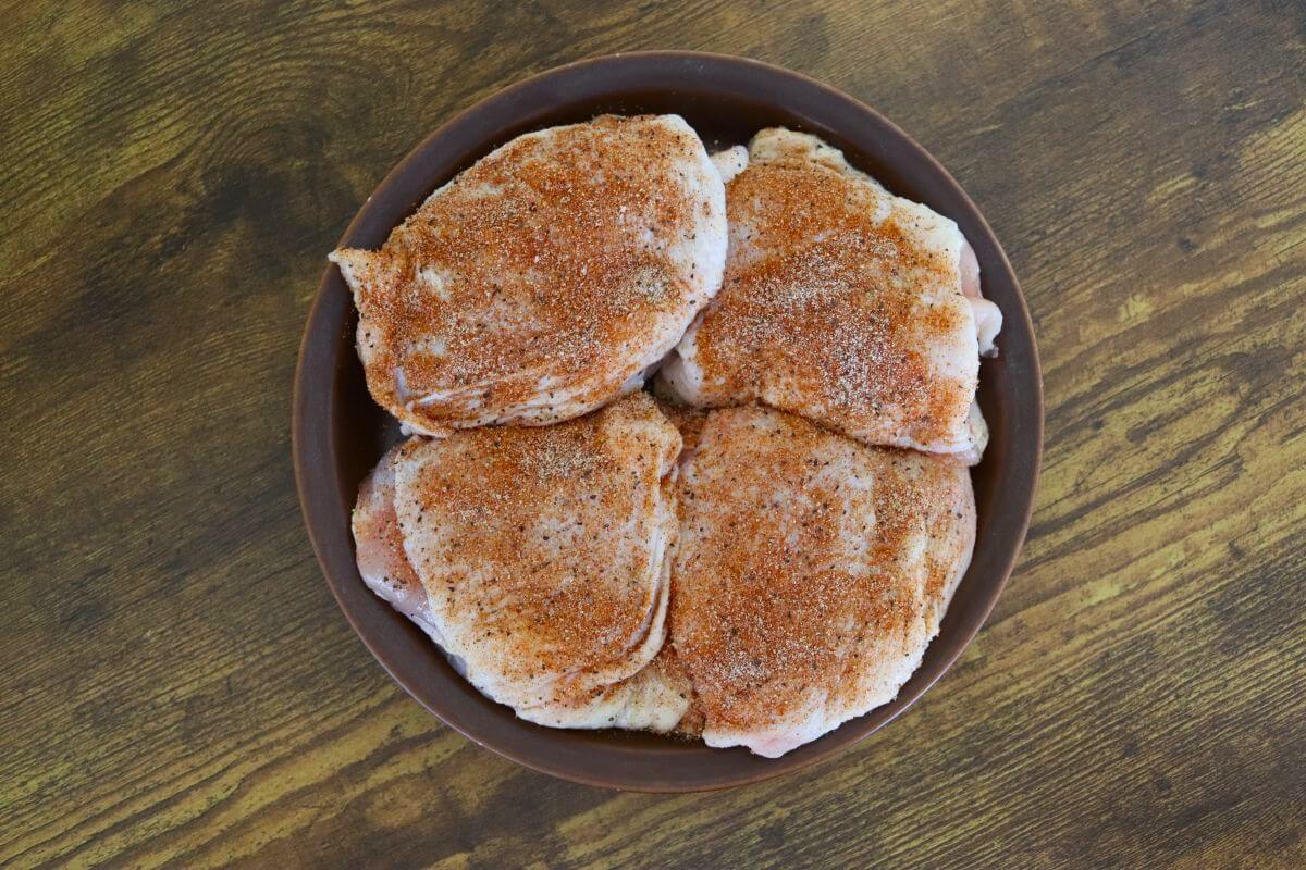 Four raw chicken thighs coated with seasoning on a brown plate on a wooden surface.