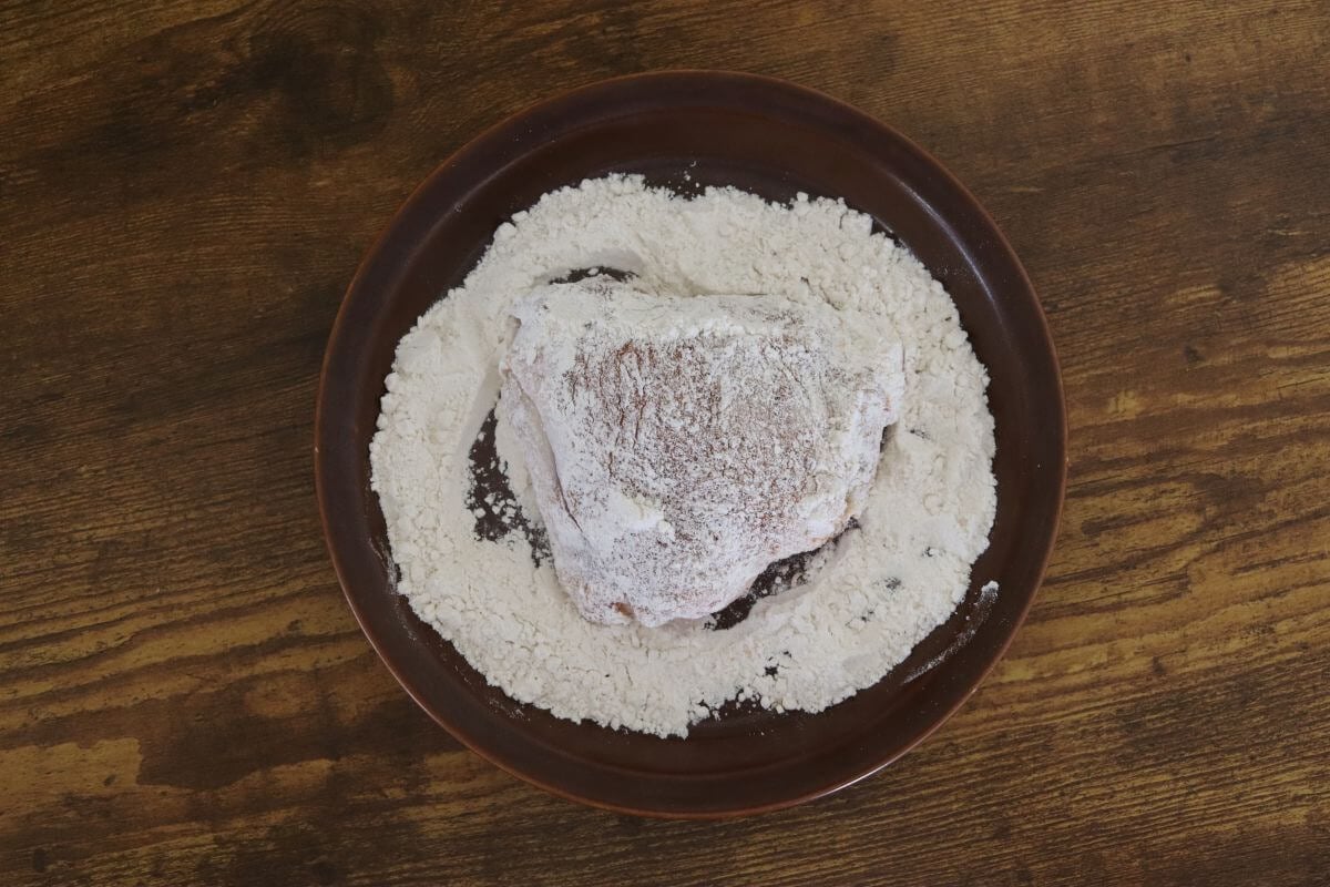 Seasoned chicken thigh dredged in flour on a brown plate over a wooden surface.