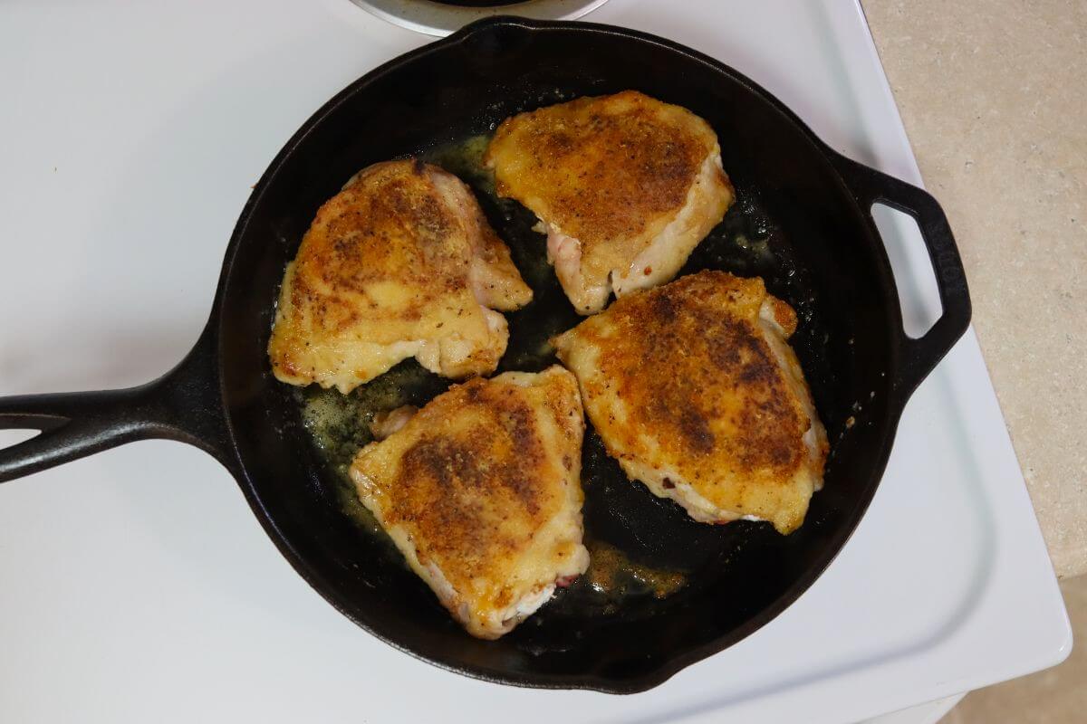 Four chicken thighs searing in a cast iron skillet on a stovetop.
