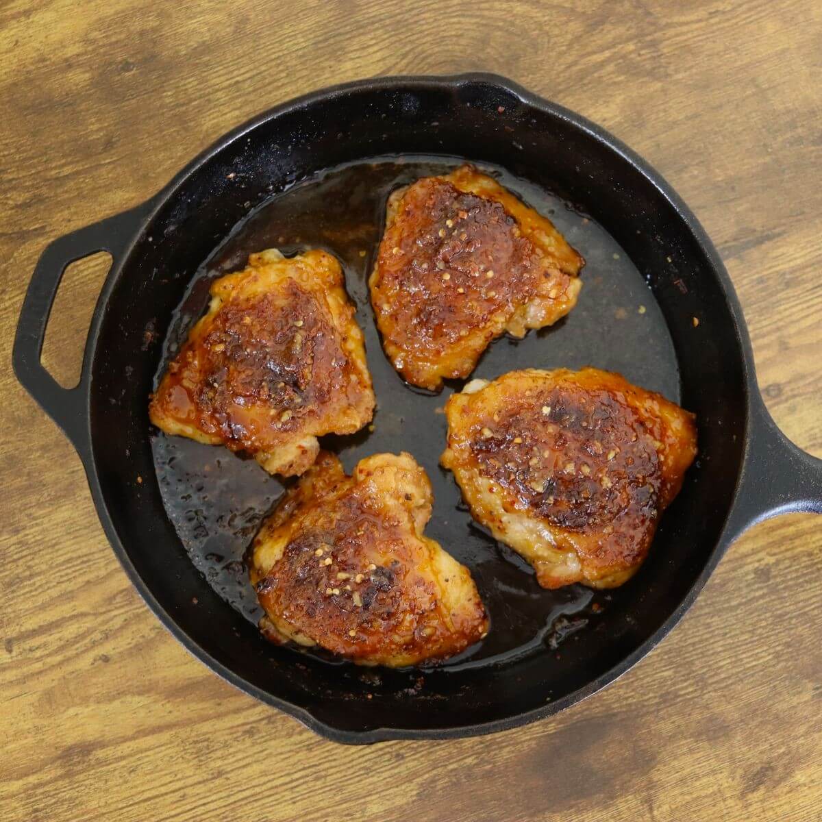 Four hot honey chicken thighs in a cast iron skillet on a wooden surface.