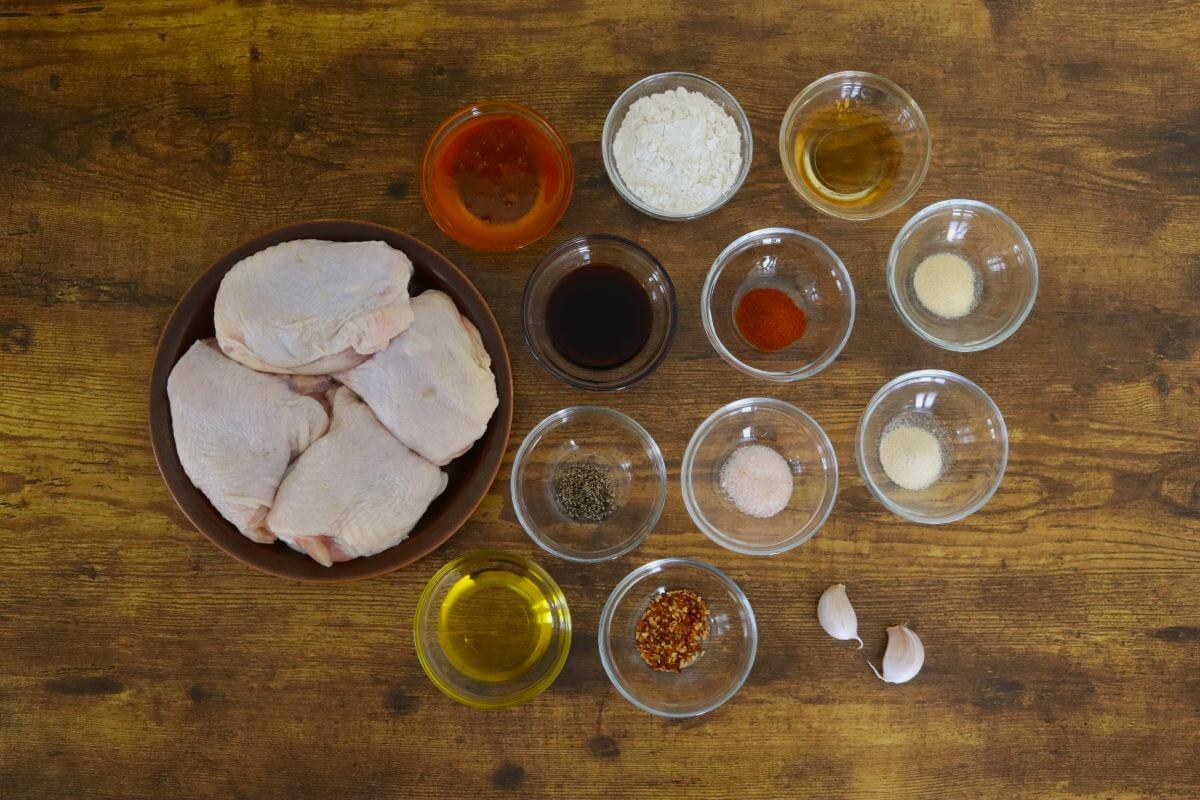 Raw chicken thighs in a bowl surrounded by small bowls of spices, oil, honey, soy sauce, and garlic on a wooden table.