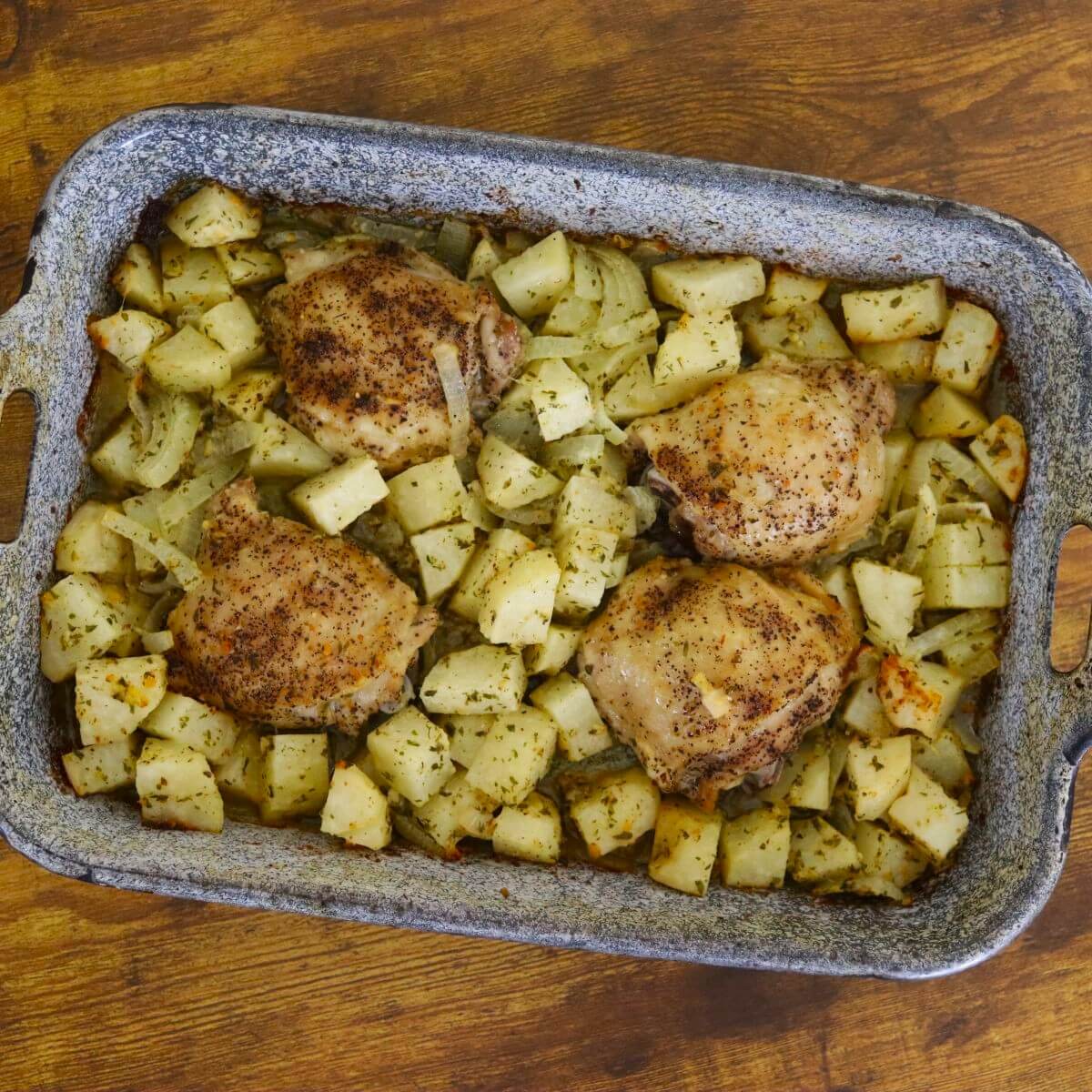 Four baked chicken thighs surrounded by roasted diced potatoes and onions in a gray rectangular baking dish.