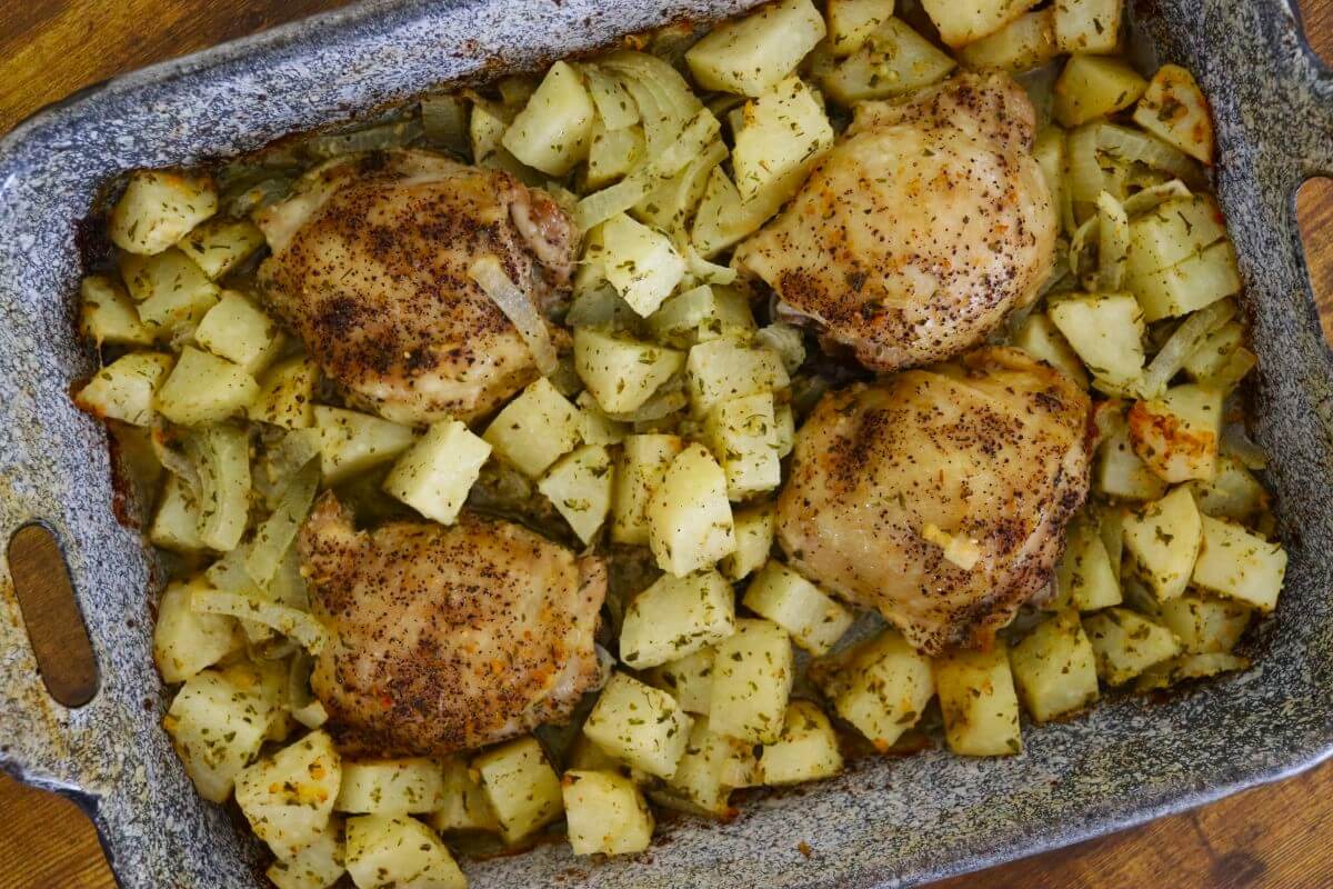 Italian baked chicken thighs with diced potatoes and sliced onions roasted together in a rectangular baking dish.