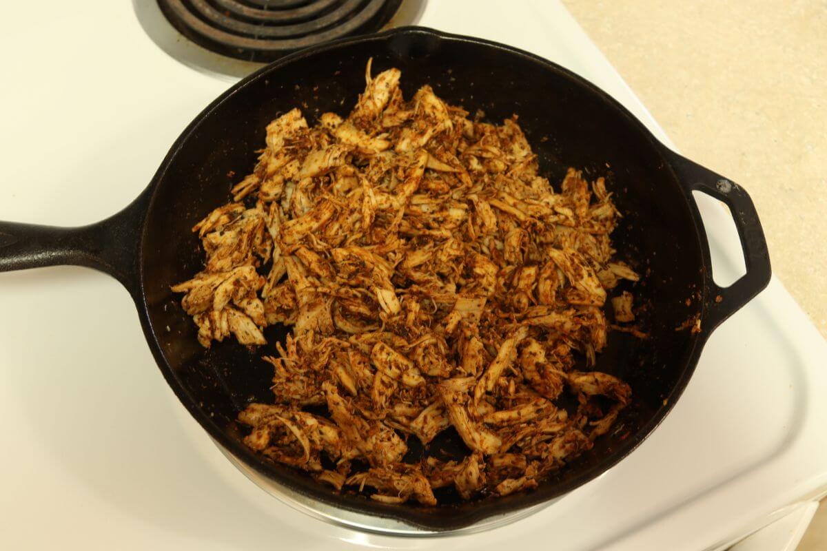 Shredded chicken coated with seasonings in cast iron skillet on stovetop.