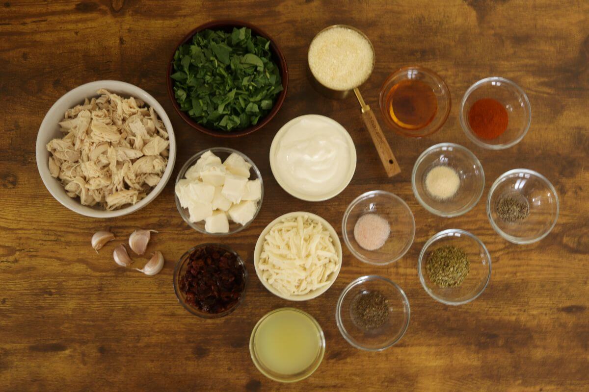 Shredded chicken, cheeses, spinach, sun-dried tomatoes, broth, and seasonings arranged on a wooden table.
