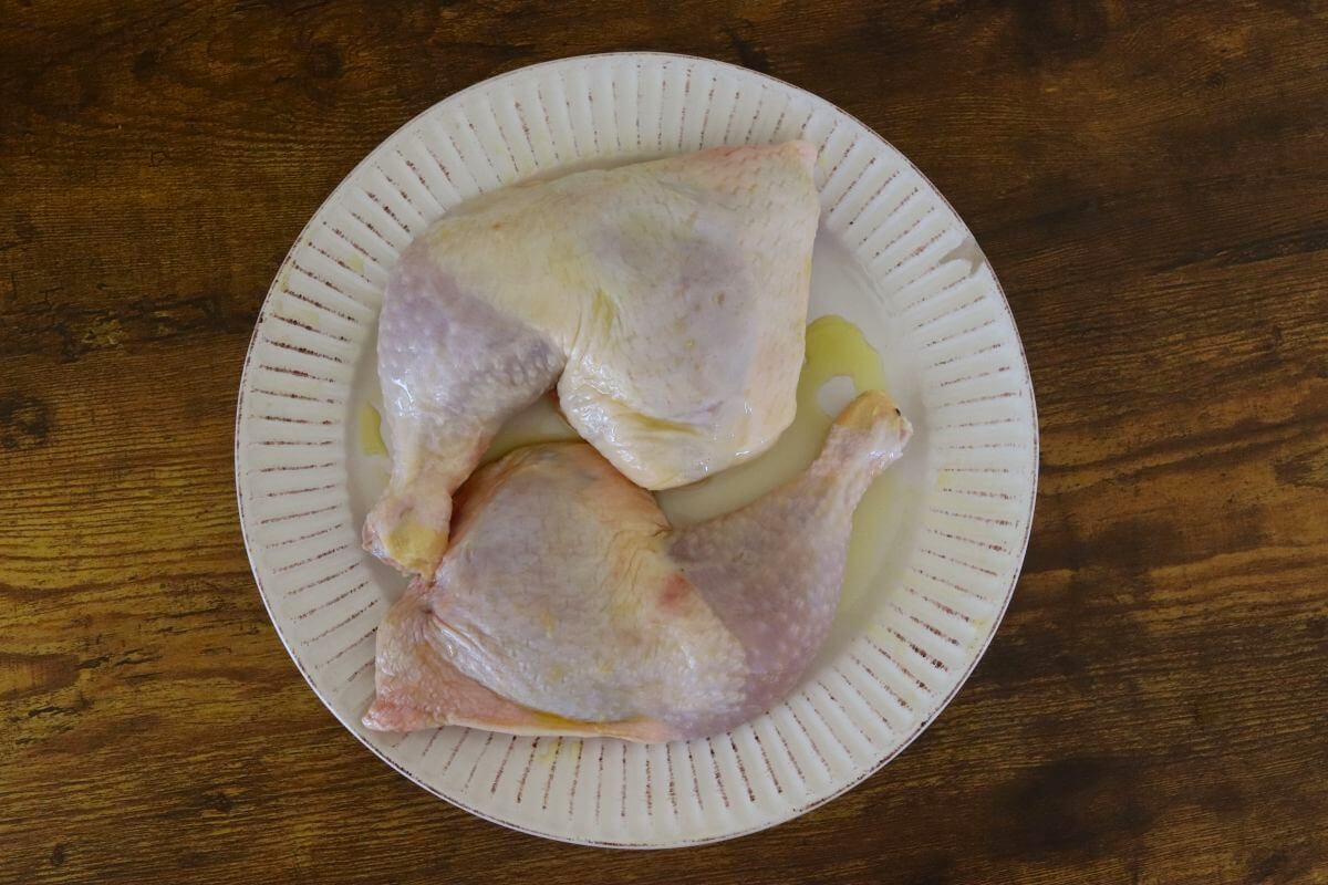 Raw chicken leg quarters lightly coated with olive oil on a white plate.