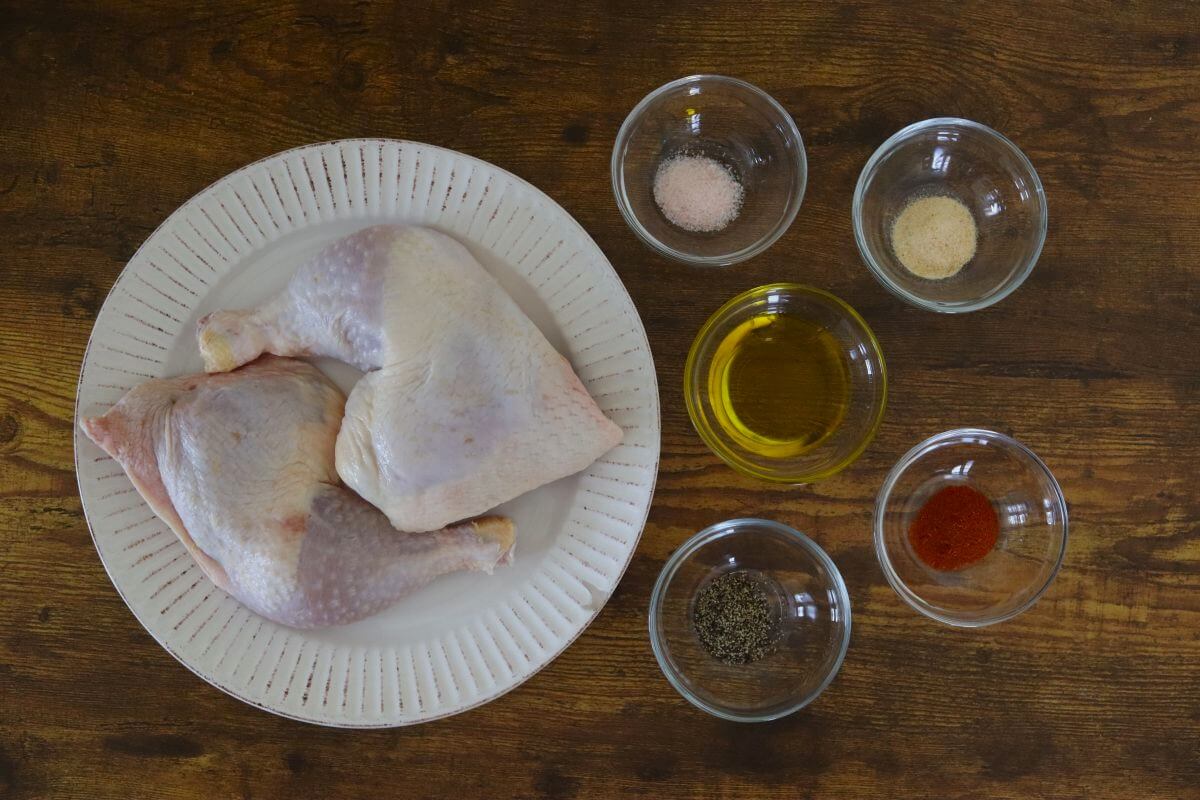Chicken leg quarters with salt, pepper, granulated garlic, smoked paprika, and olive oil in small dishes on a wooden surface.