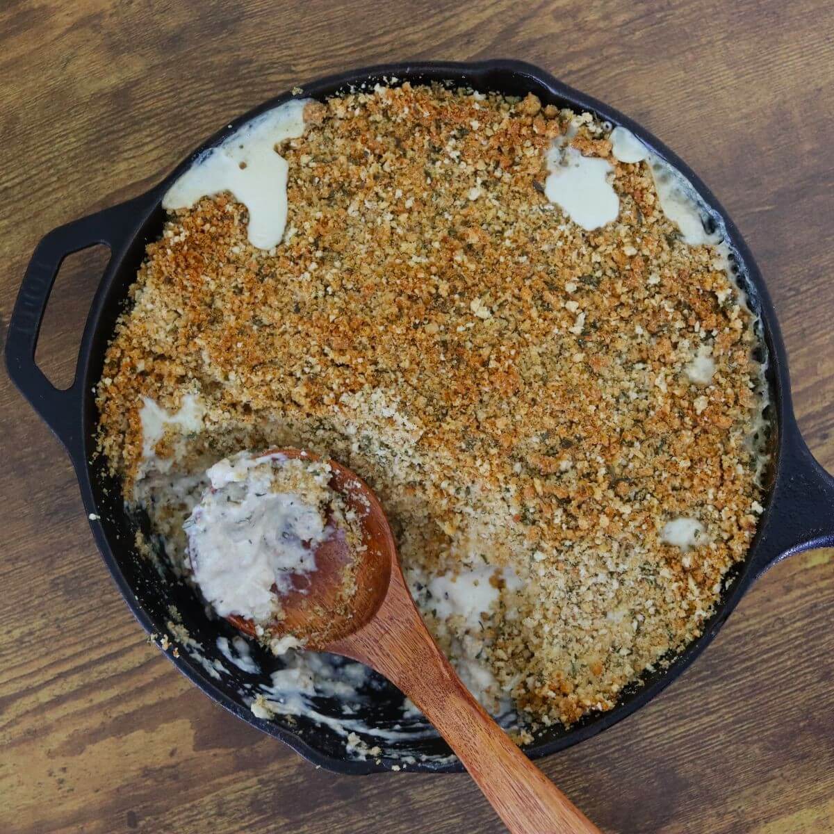 Cast iron skillet with a browned breadcrumb topping and a wooden spoon resting inside, revealing a pale mixture beneath the crumbs.