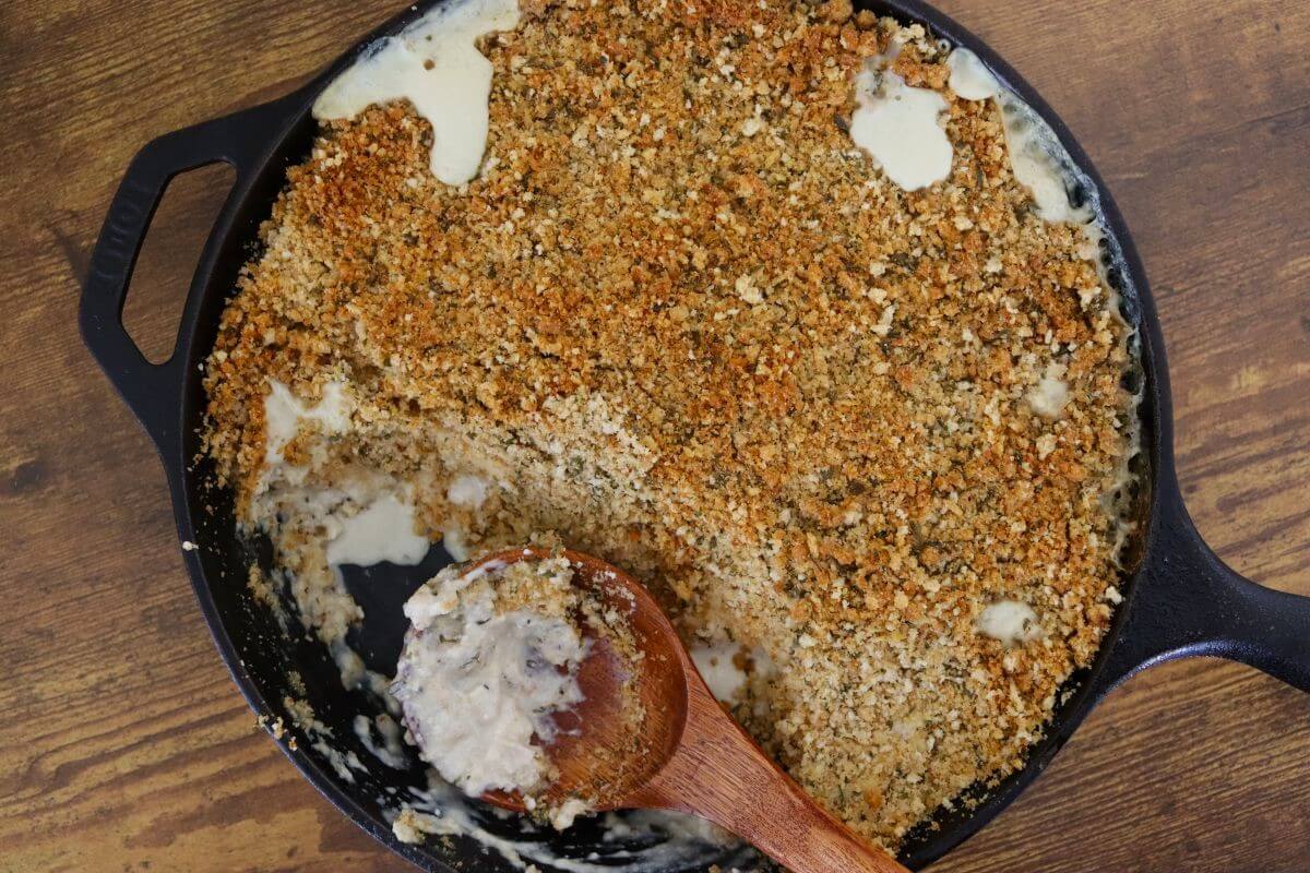 Cast iron skillet filled with a pale baked mixture topped with browned breadcrumbs, with a wooden spoon scooping from the skillet.