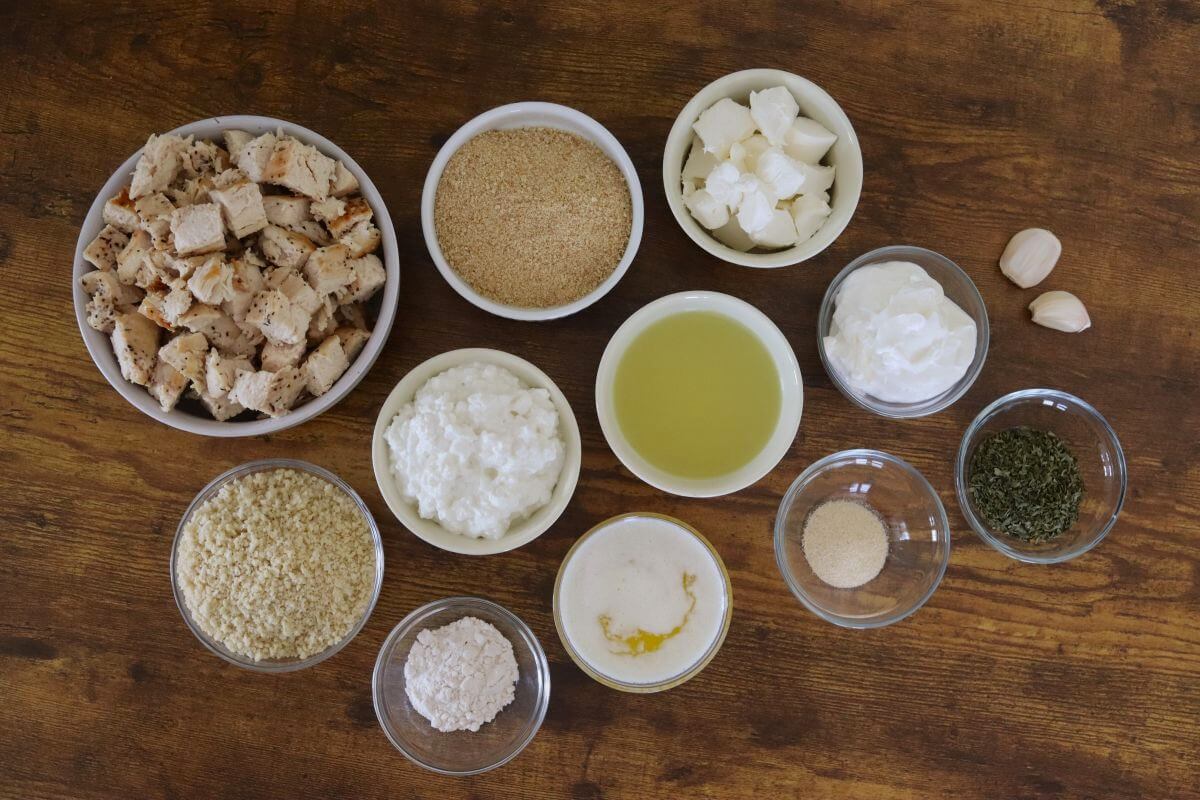 Bowls of cooked diced chicken, breadcrumbs, cream cheese, cottage cheese, sour cream, broth, flour, seasonings, and garlic arranged on a wooden surface.