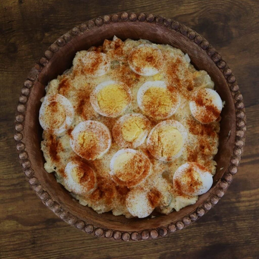Old-fashioned potato salad topped with sliced hard-boiled eggs and paprika in a carved wooden bowl on a wooden table.