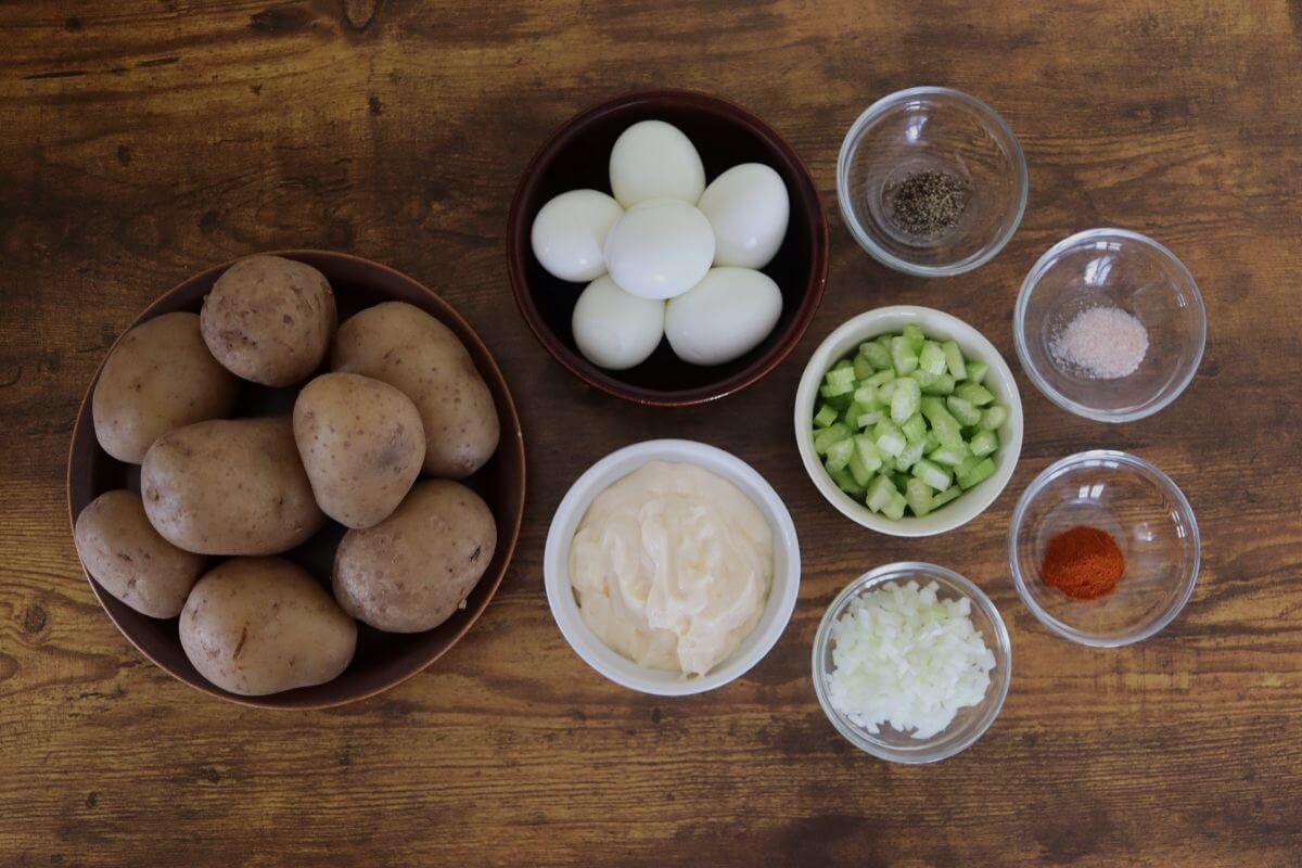 Whole potatoes, hard-boiled eggs, mayonnaise, chopped celery, diced onion, salt, pepper, and paprika on wooden table.