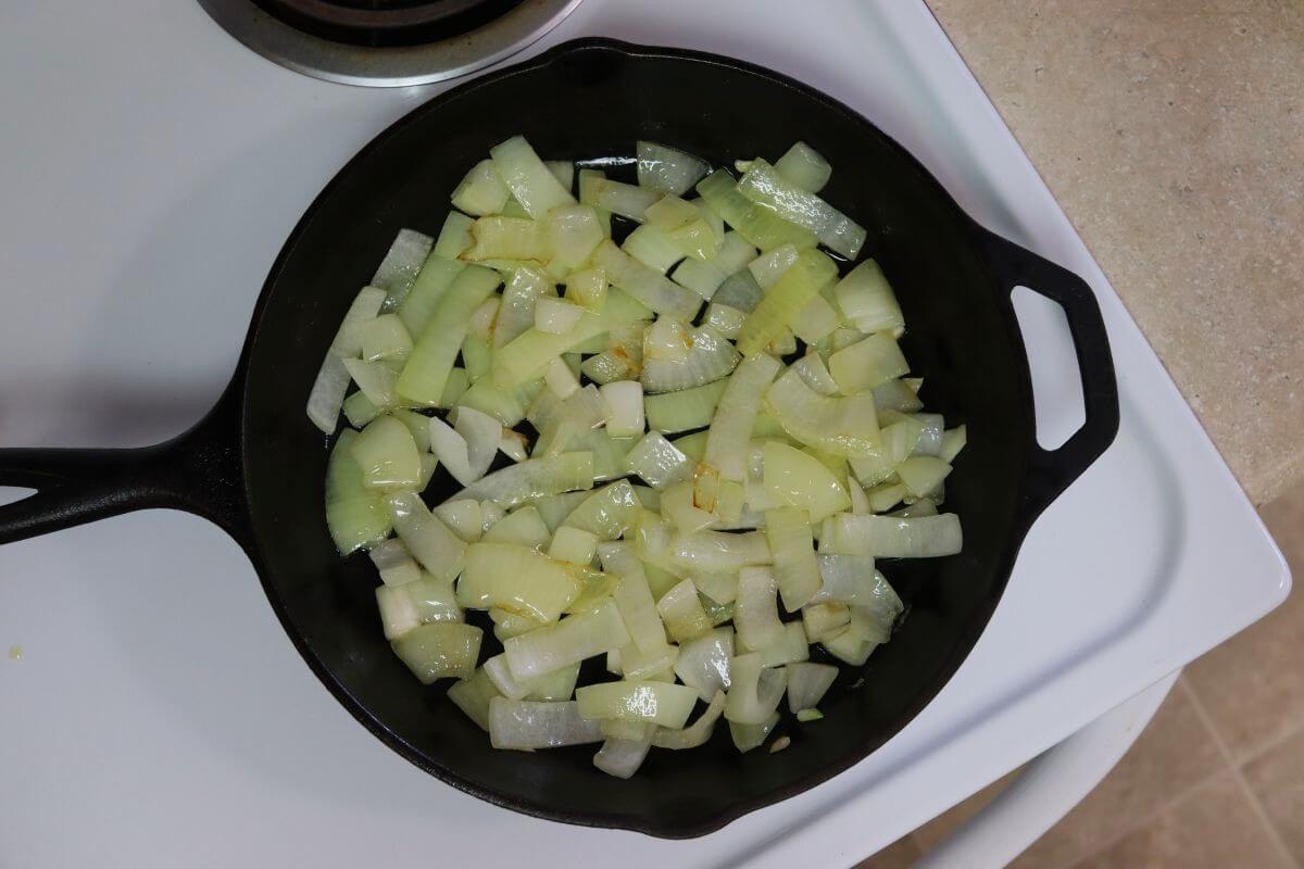 Cast iron skillet with sliced onions in oil on a white stovetop, onions glossy and lightly translucent.