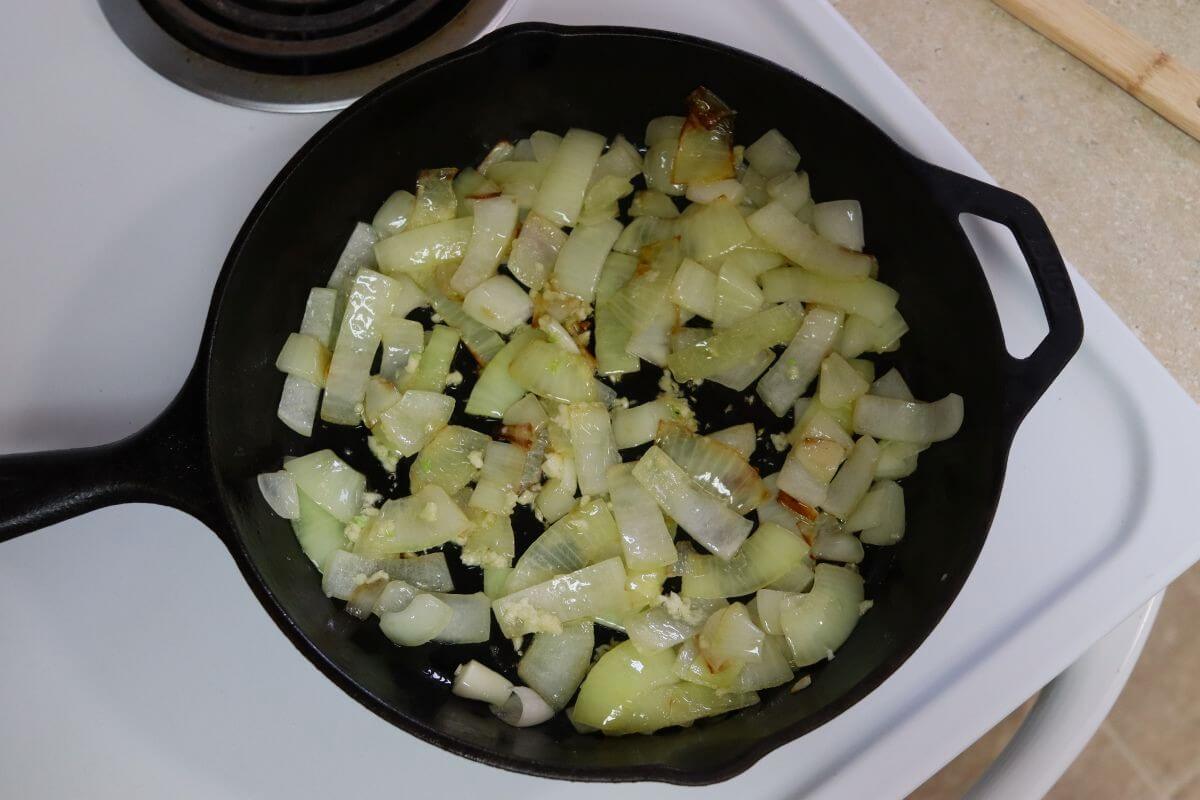 Cast iron skillet with sliced onions and minced garlic in oil on a white stovetop, onions glossy.