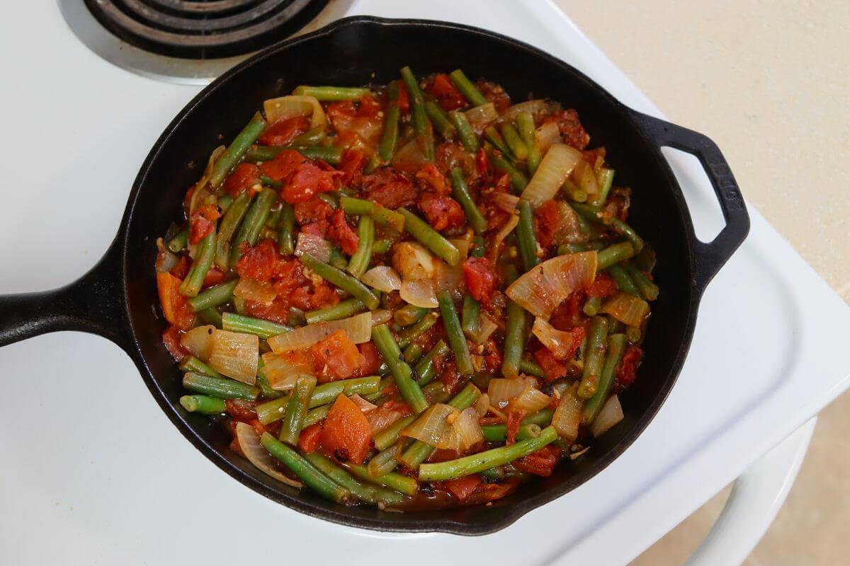 Cast iron skillet on a white stovetop filled with green beans, chopped tomatoes, and softened onion slices in a red tomato mixture.
