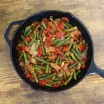 Black cast iron skillet filled with green beans, chunks of tomato, and onion pieces in a red tomato sauce on a wooden table.
