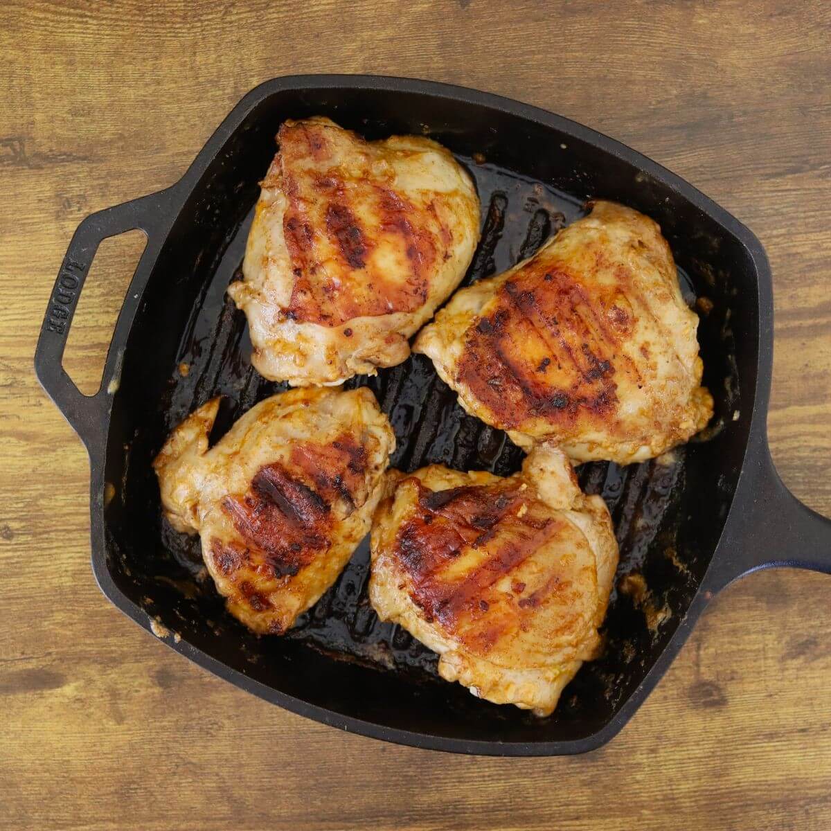 Four cooked chicken thighs resting in a cast iron grill pan, with grill ridges and rendered marinade visible.