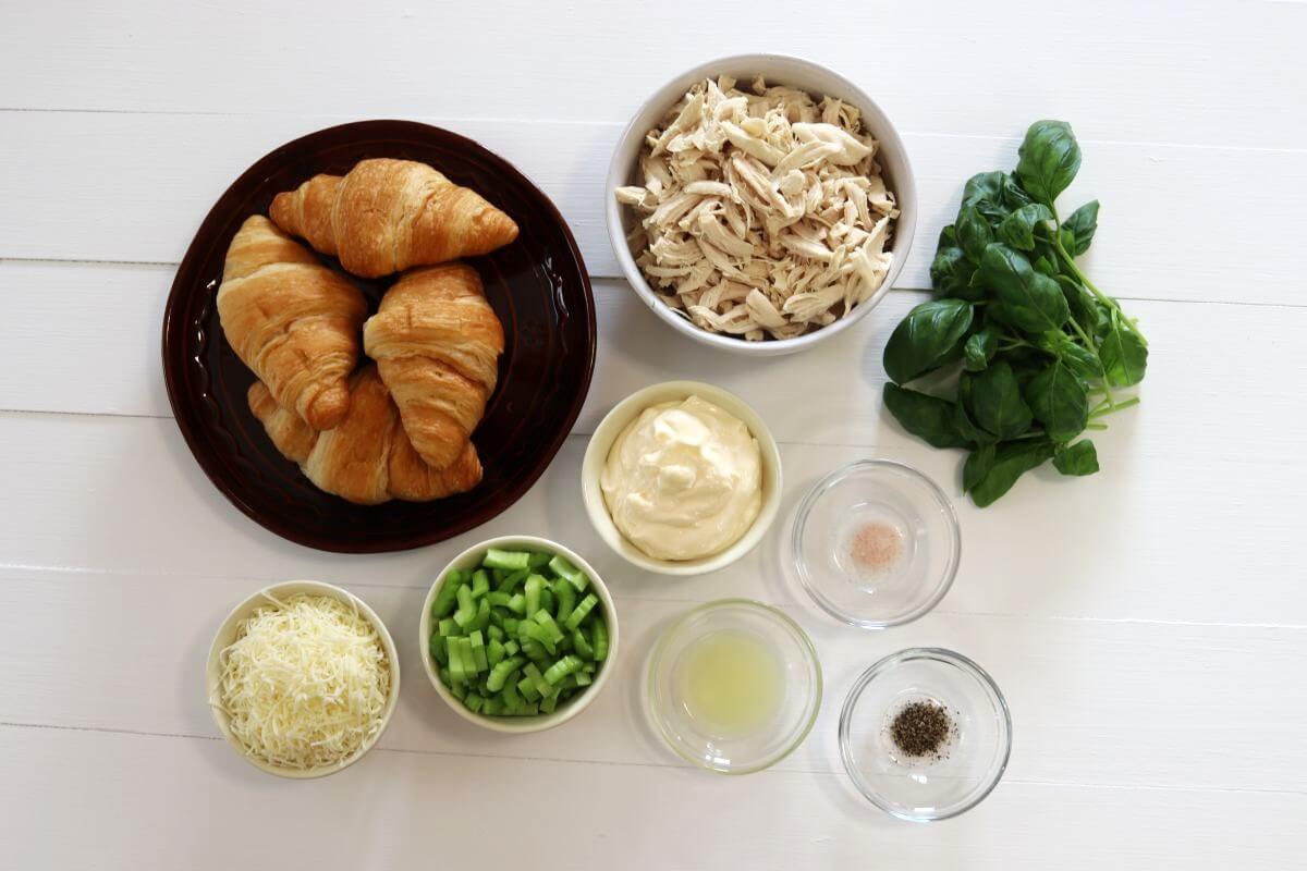 Croissants, shredded chicken, basil, celery pieces, shredded cheese, and small bowls of ingredients on a white surface.