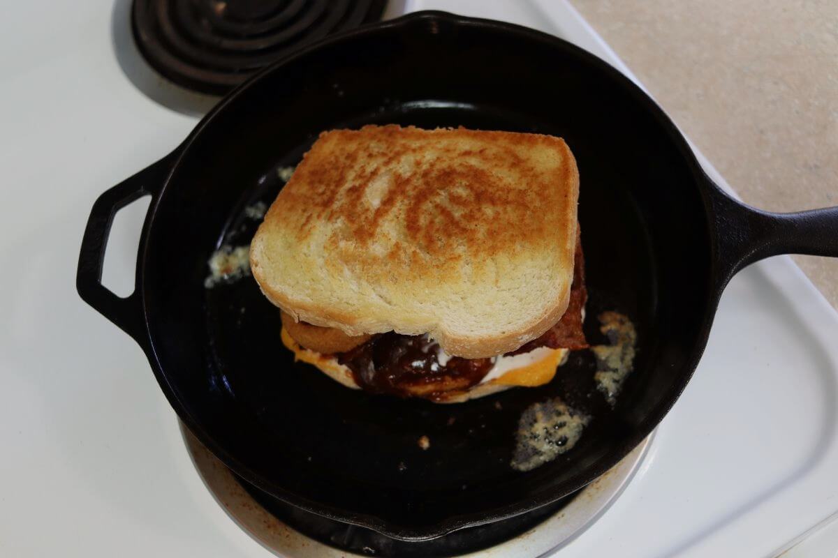 Grilled sandwich with chicken, bacon, and onion rings in cast iron skillet.
