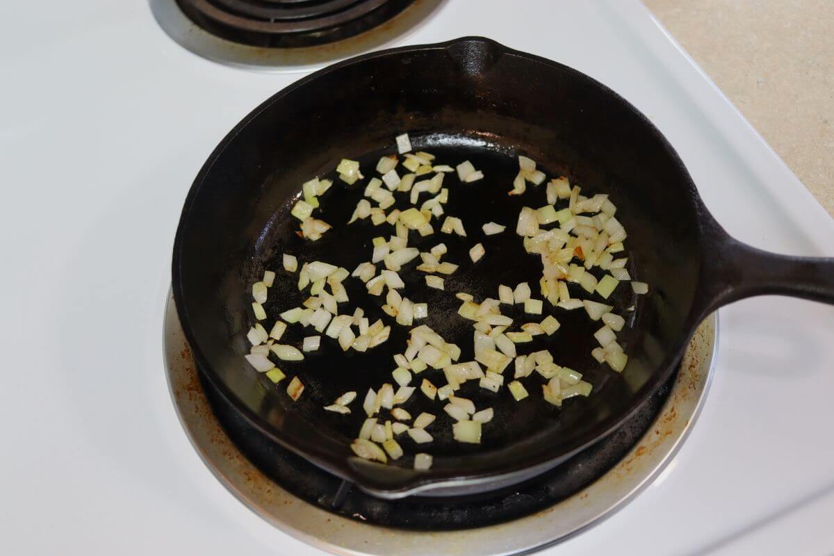 Diced onion cooking in oil in a cast iron skillet on a stovetop burner.