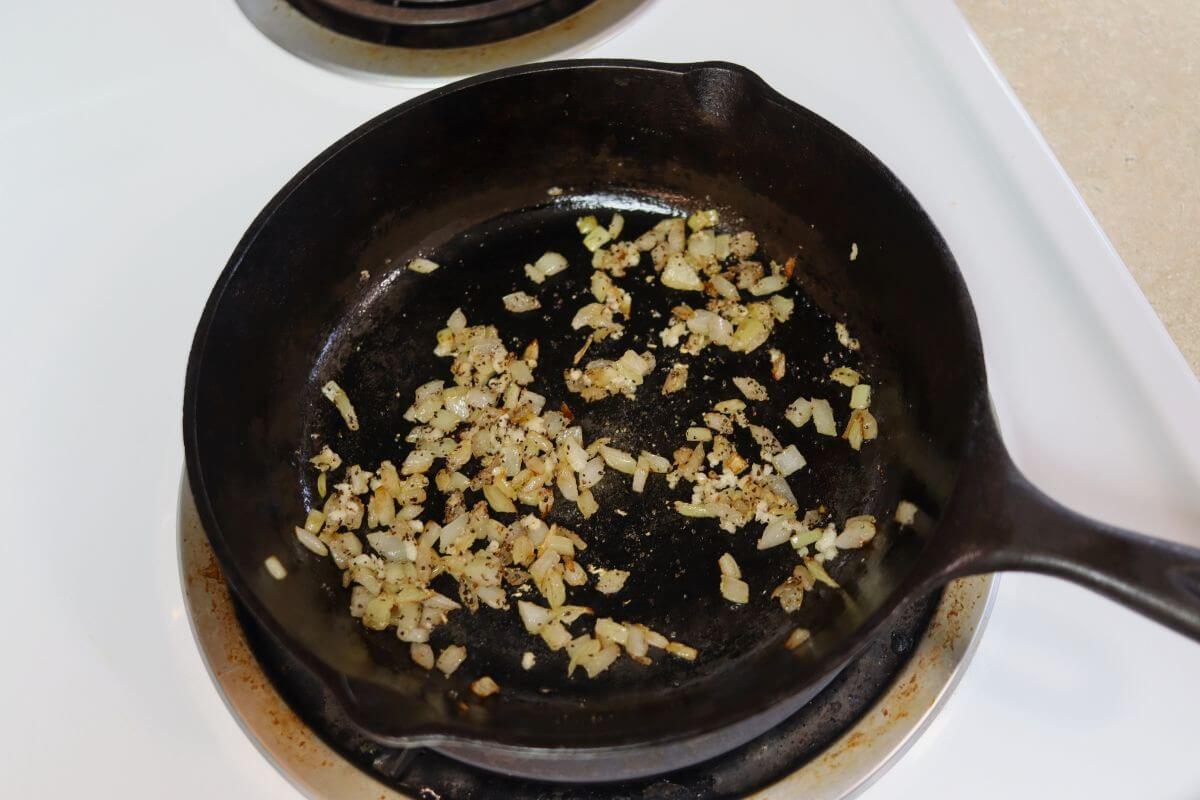 Diced onion and garlic cooking in oil with seasoning in a cast iron skillet on stovetop.