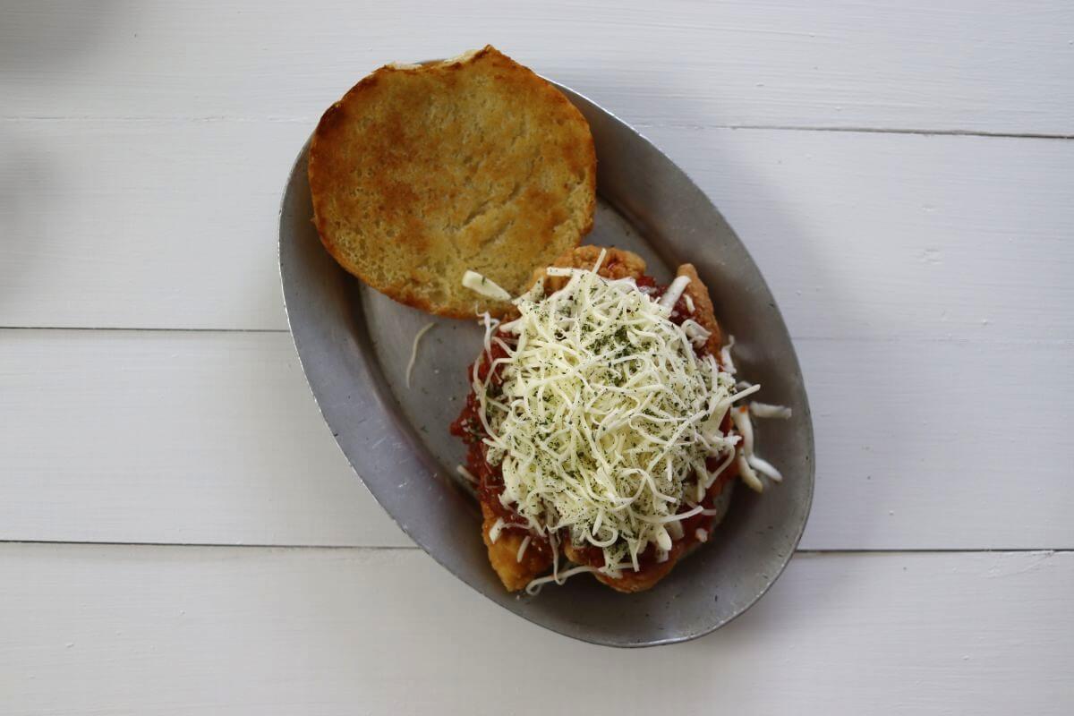 Toasted roll half topped with breaded chicken, marinara sauce, shredded cheese, and dried herbs on pan.