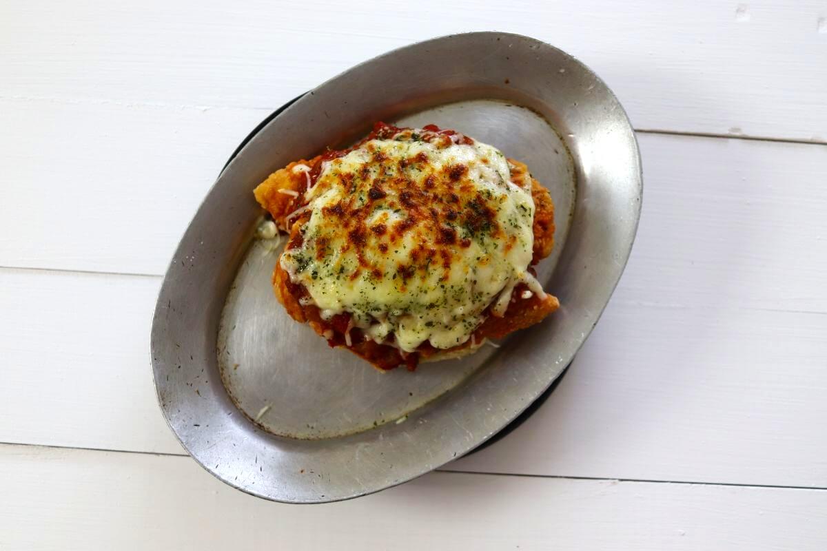Breaded chicken with marinara sauce and melted cheese on toasted roll half on metal pan.