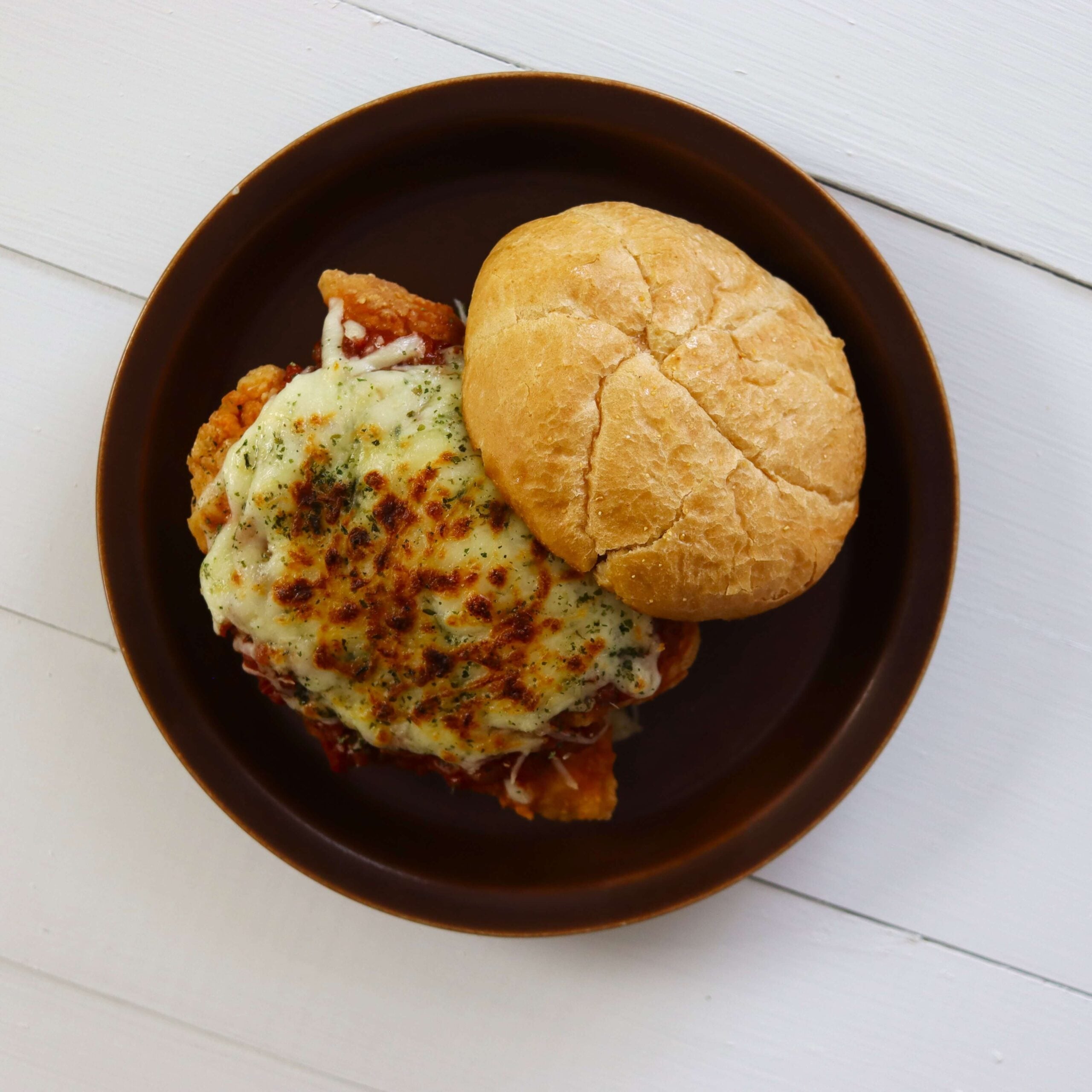 Breaded chicken with marinara and melted cheese on roll half with top bun on brown plate.