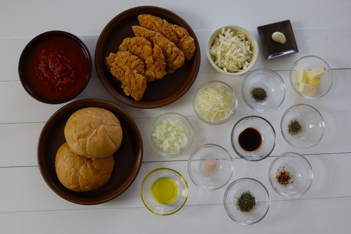 Bowls of marinara, breaded chicken tenders, sandwich rolls, shredded cheeses, chopped onion, butter, oil, and seasonings on table.