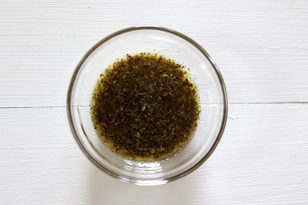 Oil mixed with dried herbs and seasonings in a glass bowl on a white surface.