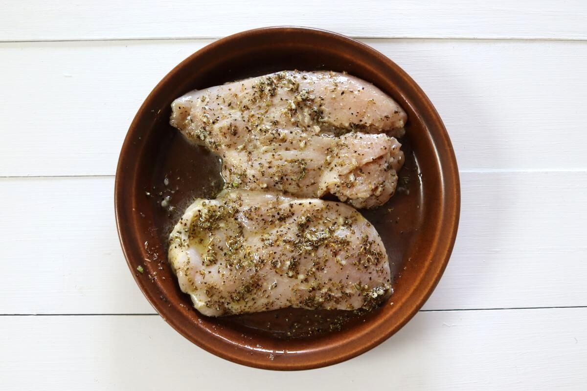 Raw chicken breasts coated with oil and dried herbs in a brown bowl.