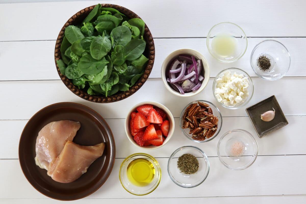 Raw chicken breasts on a plate with bowls of spinach, sliced strawberries, pecans, feta, red onion, garlic, oil, and seasonings.