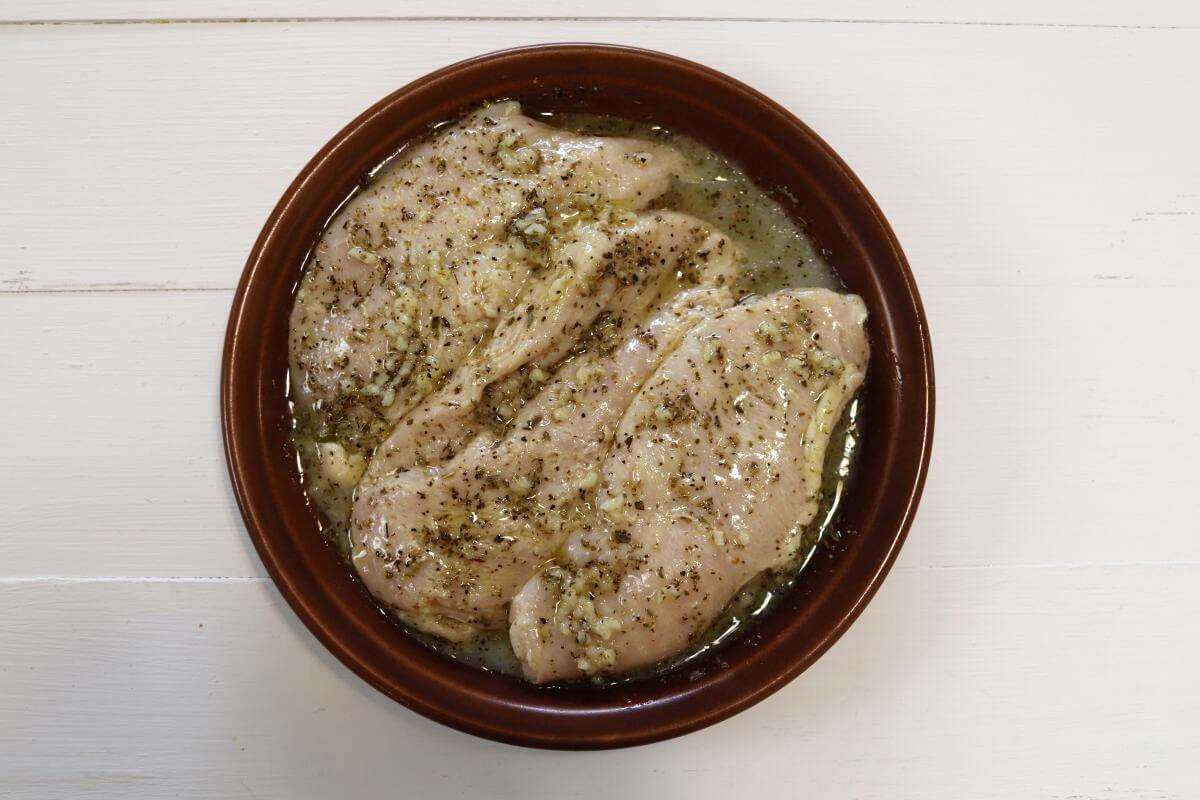 Raw chicken breasts in a bowl coated with oil, herbs, garlic, and seasoning.
