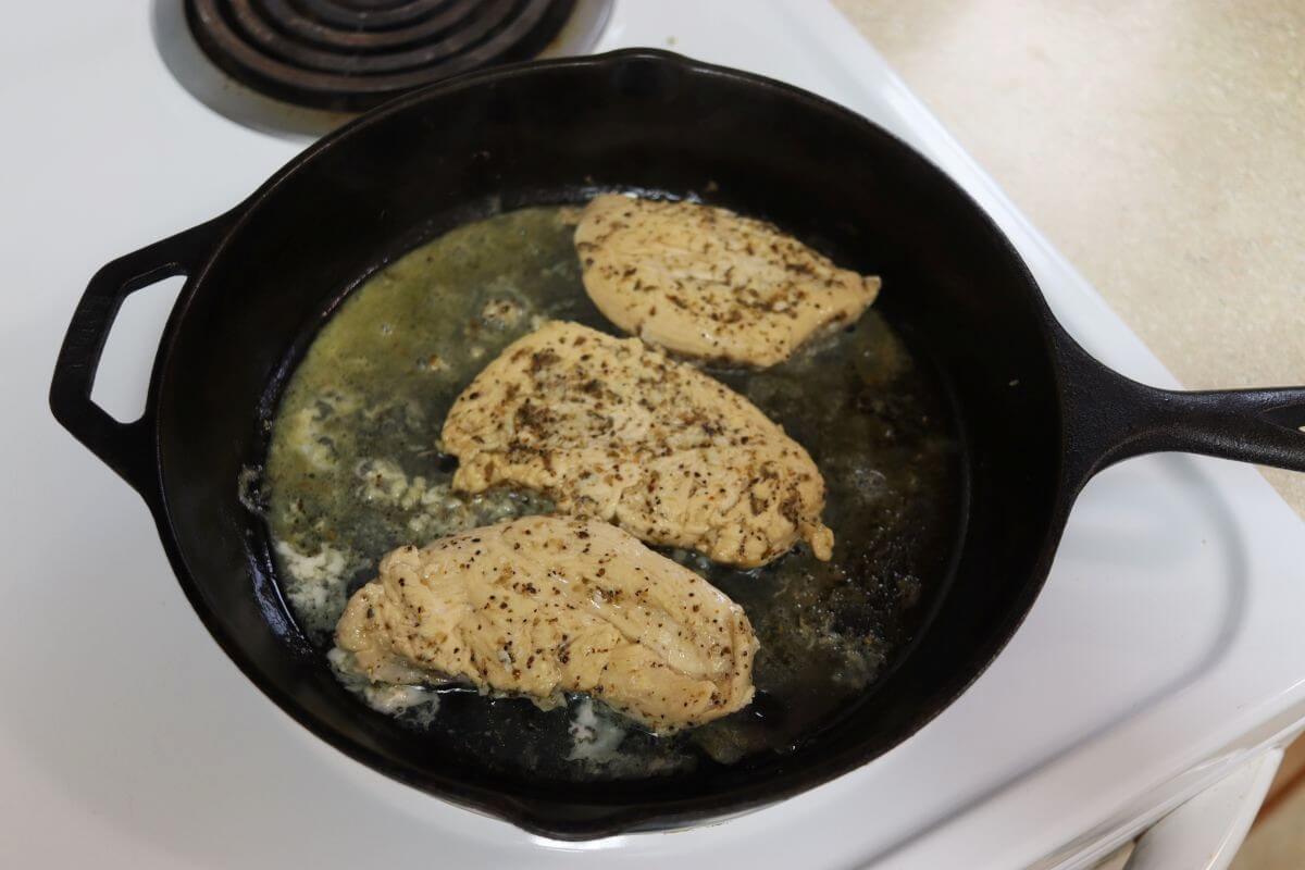 Chicken breasts cooking in a cast iron skillet with oil, garlic, and herbs on stovetop.