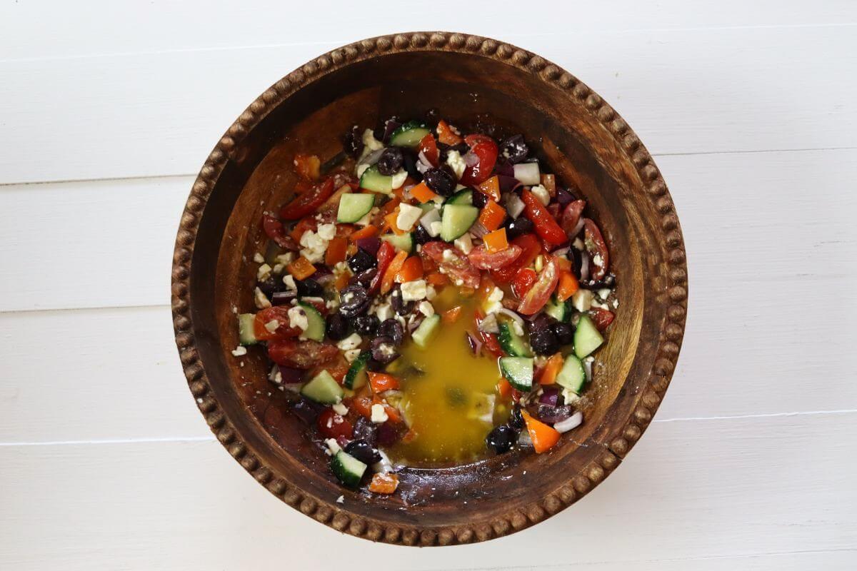 Vegetables, olives, and feta in a wooden bowl with oil and vinegar dressing added.