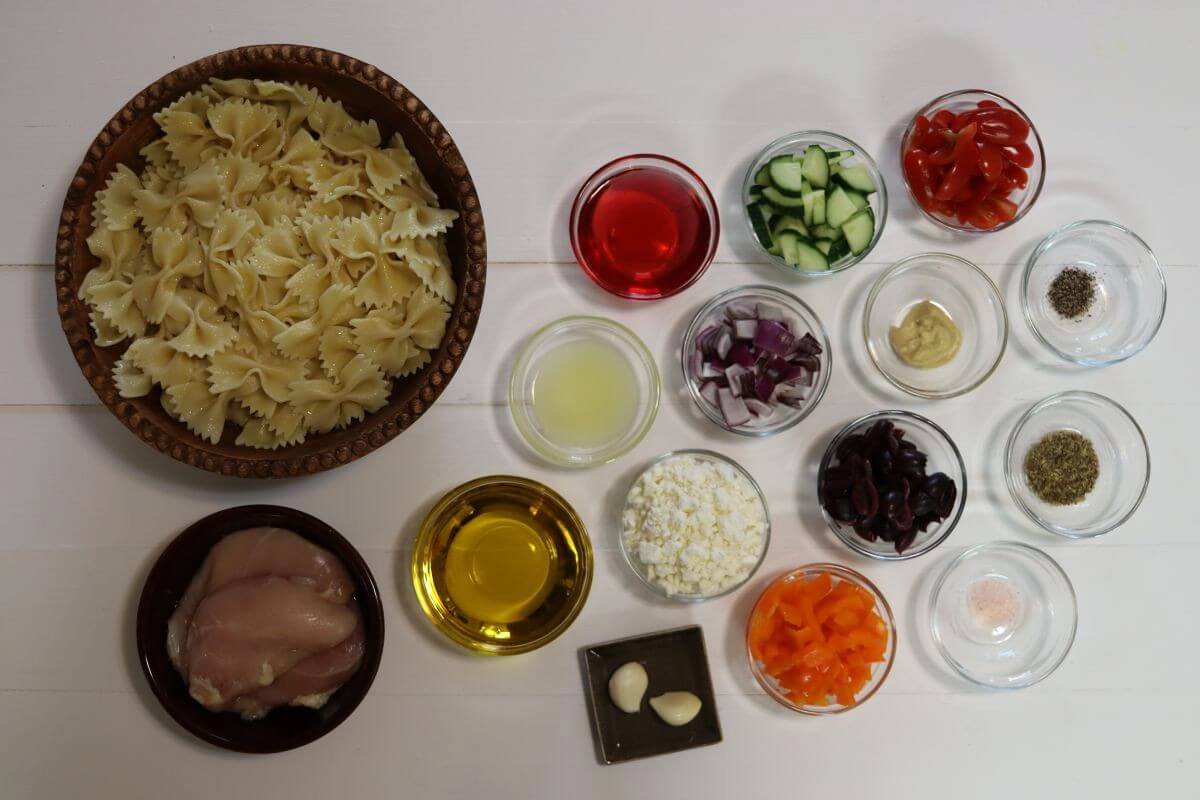 Bowl of farfalle pasta with raw chicken, chopped vegetables, feta, oil, and seasonings in small dishes.