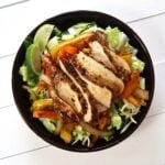 Salad bowl with romaine lettuce, grilled chicken slices, sautéed peppers and onions, shredded cheese, and lime wedges.