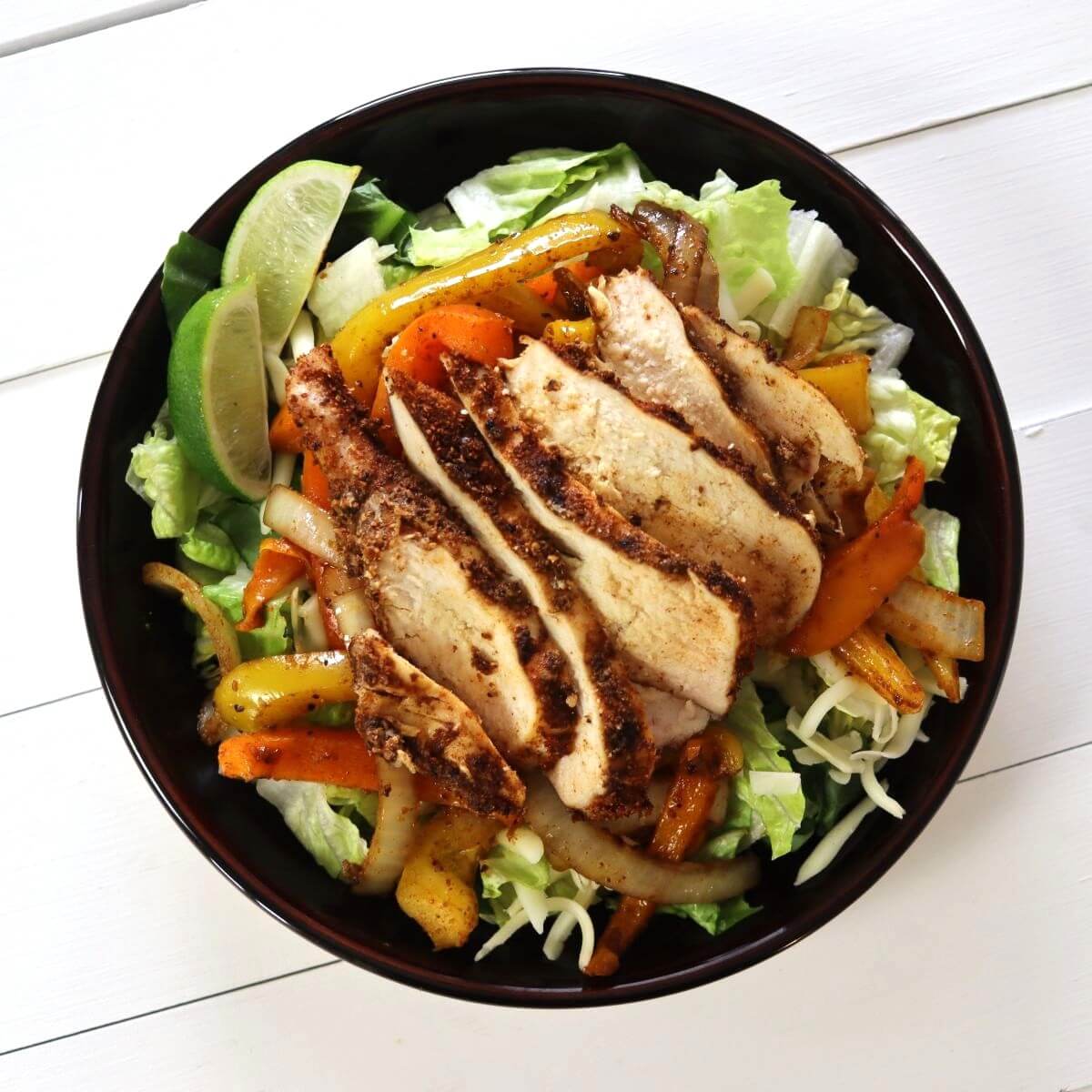 Salad bowl with romaine lettuce, grilled chicken slices, sautéed peppers and onions, shredded cheese, and lime wedges.