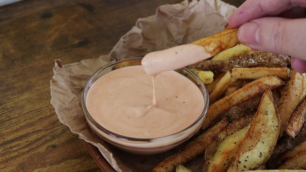 Hand dipping seasoned hand cut french fries into pink sauce in glass bowl.