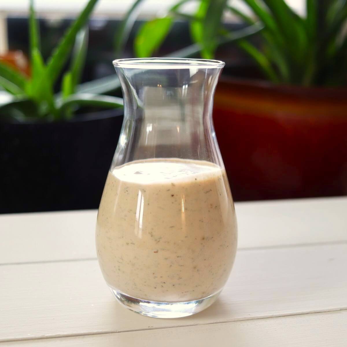 Glass carafe of herb-flecked dressing on white table with potted plants behind.