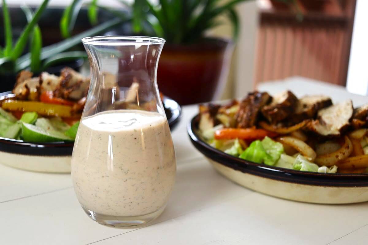 Glass carafe of creamy dressing on a white table with two chicken salads behind.