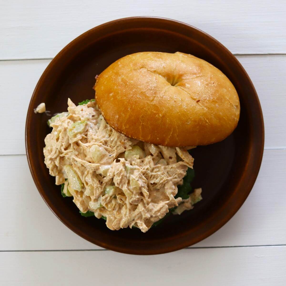 Chicken salad with celery on lettuce in a bun on a brown plate on a white wooden surface.