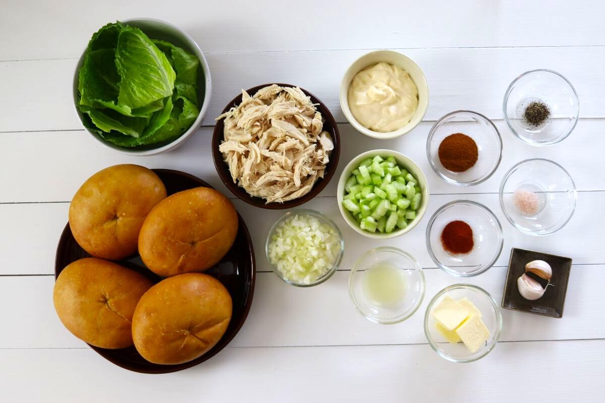 Bowls of shredded chicken, celery, onion, mayonnaise, spices, lettuce, buns, garlic, butter, and lemon juice.