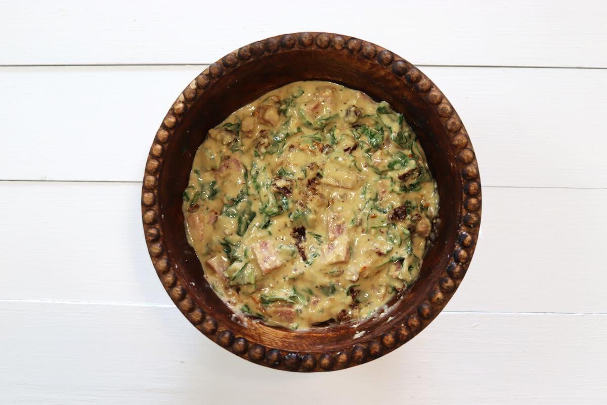 Salami, sun-dried tomatoes, and spinach mixed with creamy dressing in wooden bowl on white wooden surface.